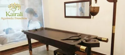 Logotipo de Kairali Ayurveda Franchise sobre una sala de tratamiento con camilla de masaje de madera y recipiente de aceite suspendido