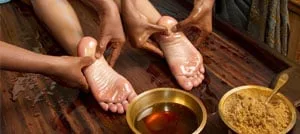 Terapisti che massaggiano olio caldo sui piedi di un paziente, accanto a ciotole di ottone con olio e polvere di erbe