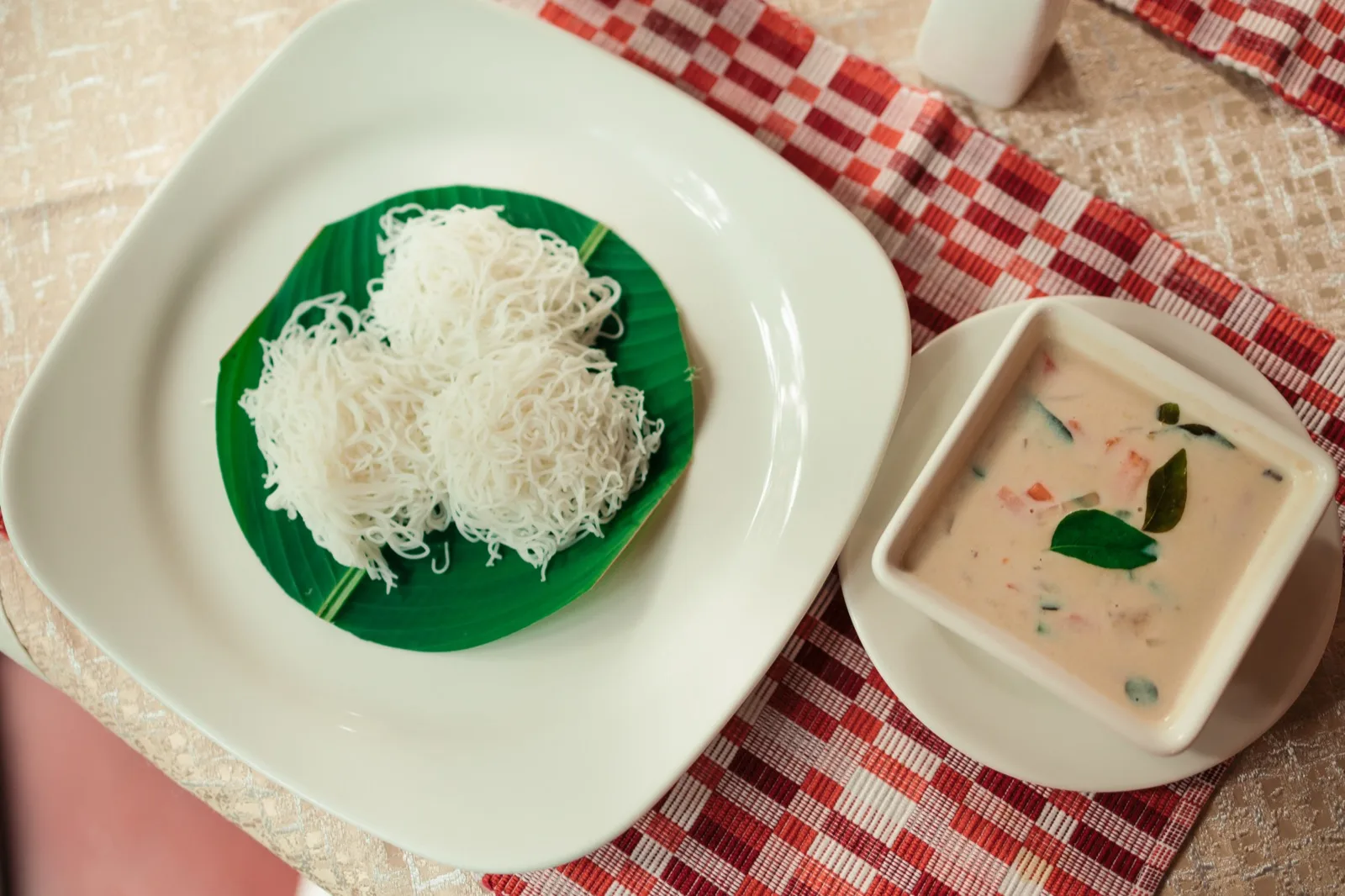 俯拍：白盘中的芭蕉叶上盛着 idiyappam（米线饼），旁边是一方碗蔬菜