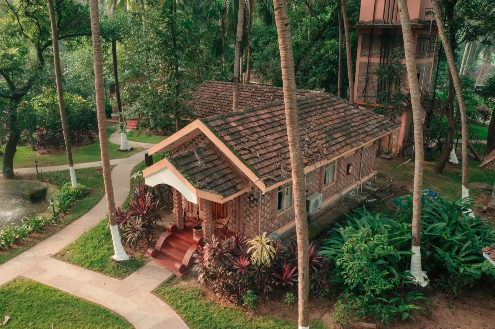 俯瞰一座瓦顶砖砌小屋，带拱形门廊和红色台阶；四周是刷白的棕榈树干