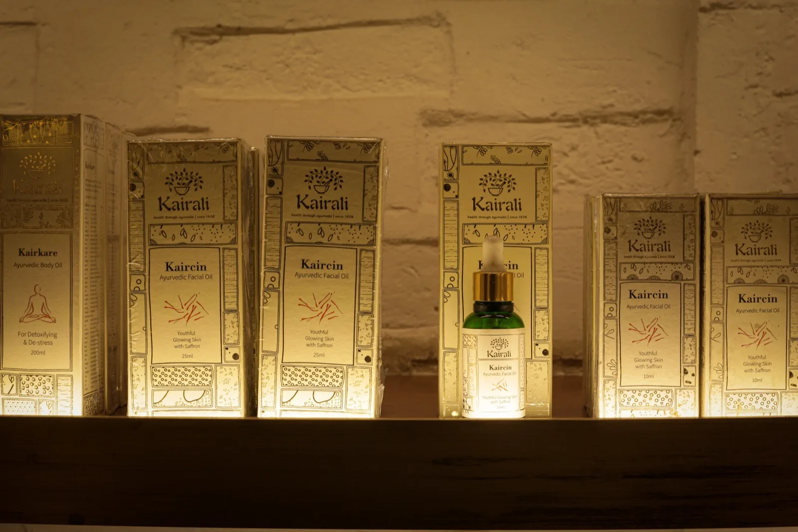 رفّ لعرض منتجات Kairali مع علب مضاءة من الخلف لزيت الوجه الأيورفيدي Kaircin Ayurvedic Facial Oil وزيت الجسم Kairkare Body Oil، إضافةً إلى قطّارة خضراء