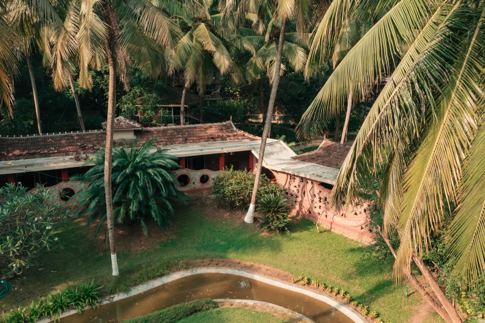 Veduta dall'alto, attraverso le fronde delle palme, di edifici in laterite con tetti di tegole e decorazioni in mattoni; sotto, un prato e un canale d'acqua sinuoso