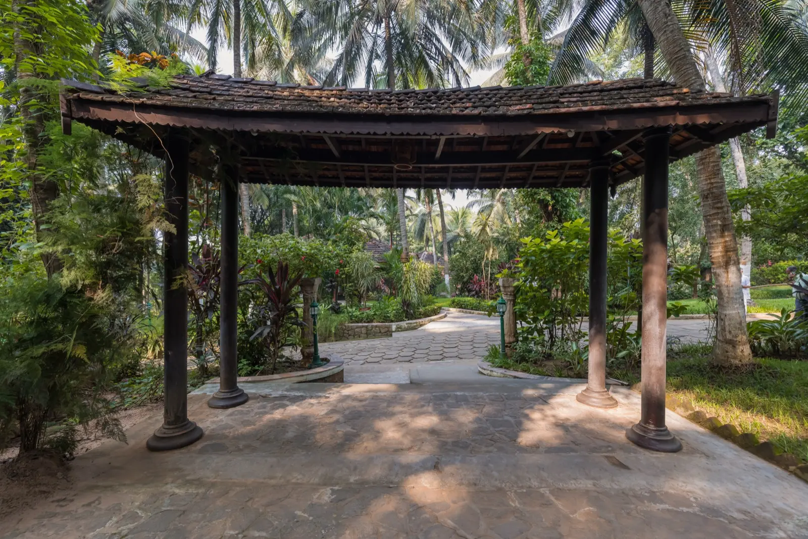 Gazebo aperto in legno con tetto curvo di tegole su colonne in legno scuro; vista verso il cortile con pavimentazione esagonale e i vialetti del giardino più avanti