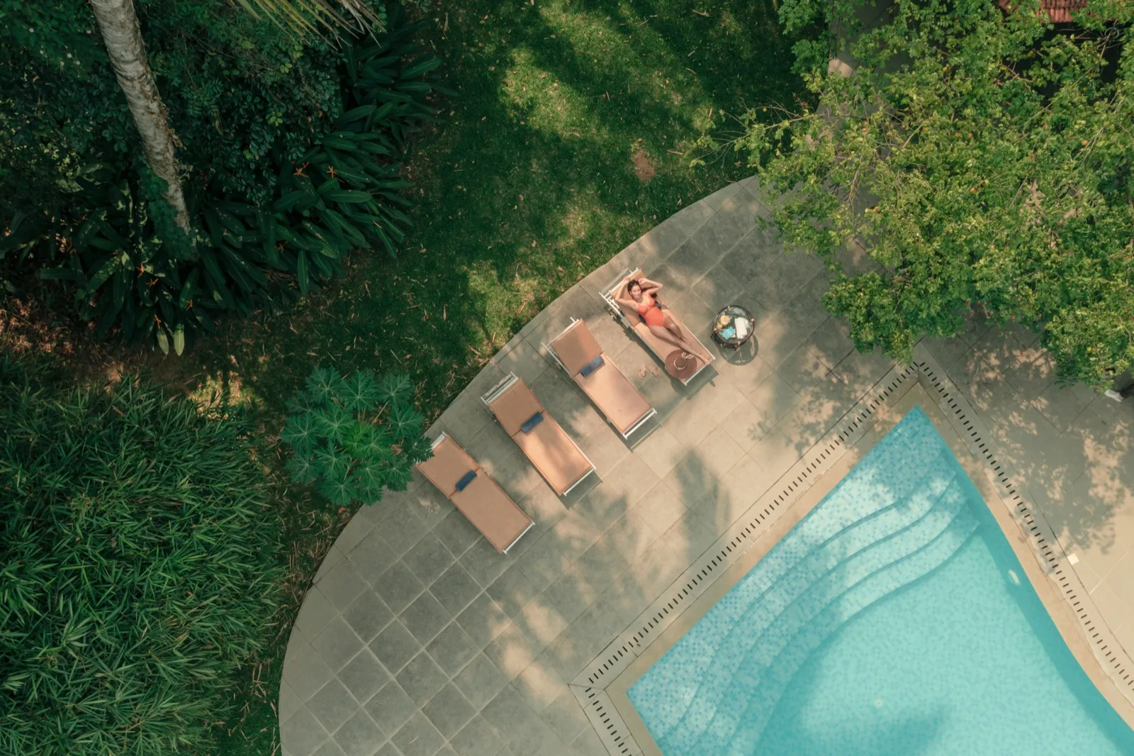 Ripresa zenitale con drone del bordo piscina con una donna distesa su un lettino accanto a un tavolino