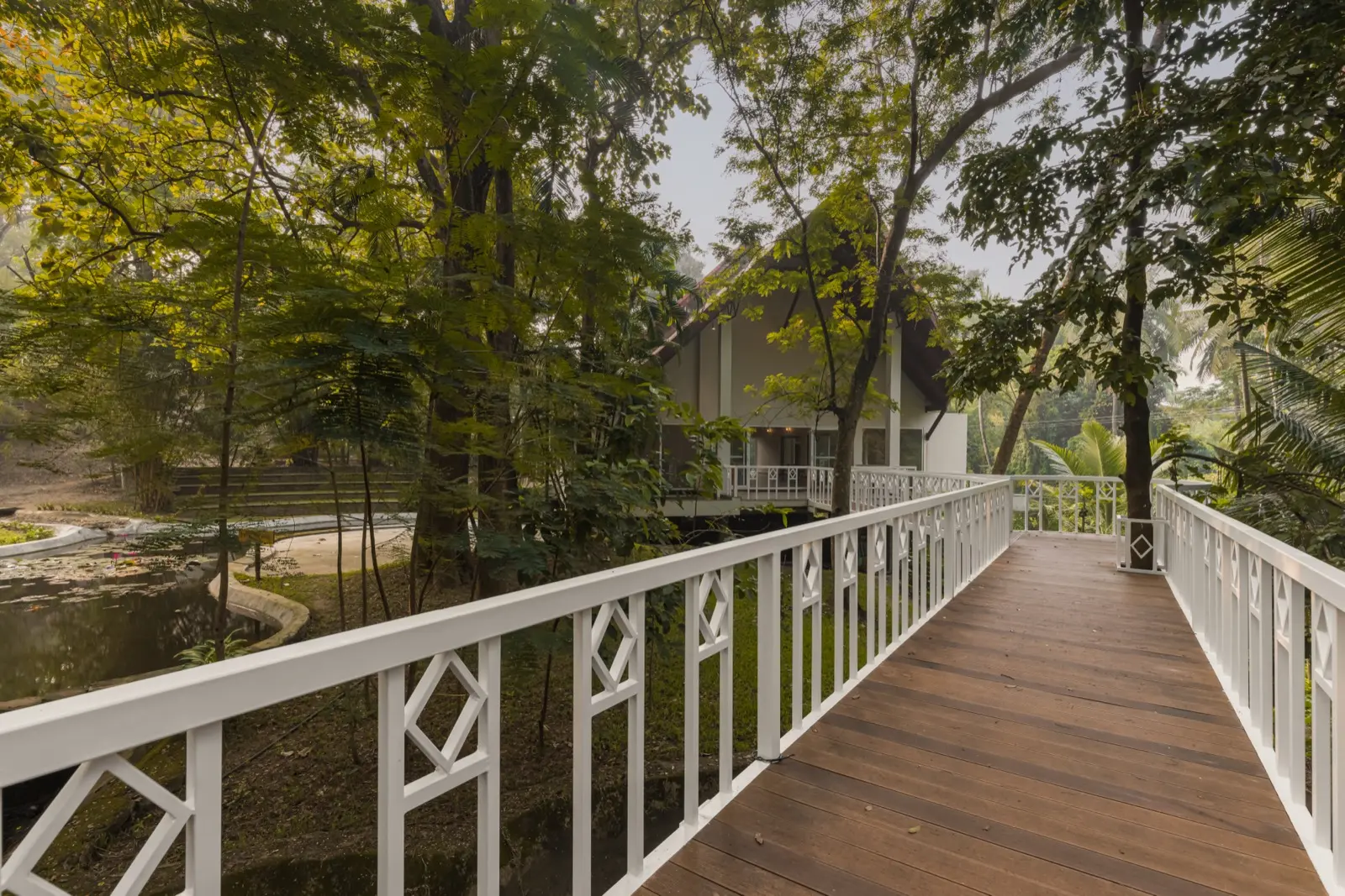 Passerella in legno con ringhiere bianche che attraversa uno stagno di loti verso un edificio con tetto a capanna tra alberi alti; a sinistra si vedono le gradinate dell'anfiteatro