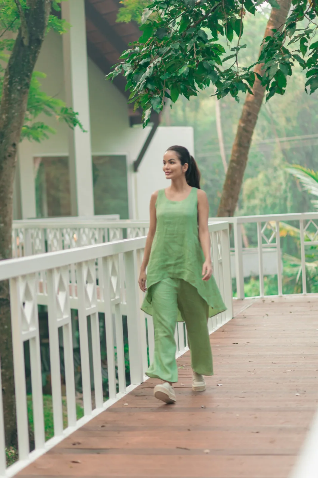Donna in abito di lino verde colta a metà passo su una passerella in legno con ringhiera bianca; alle spalle un cottage e alberi tropicali