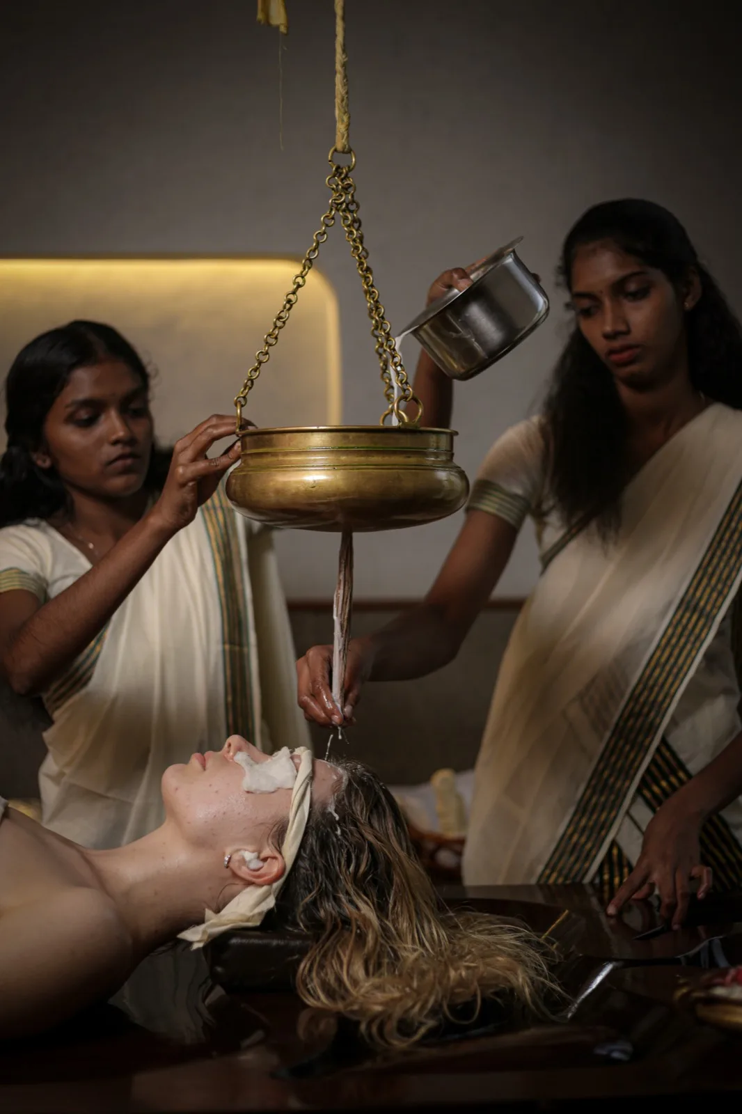 タクラダーラ（Thakaradhara）：タクラダーラ（Thakradhara）の施術中。セラピストの一人が吊り下げた真鍮の容器を支え、もう一人が薬用の液体を注ぎながら誘導しています