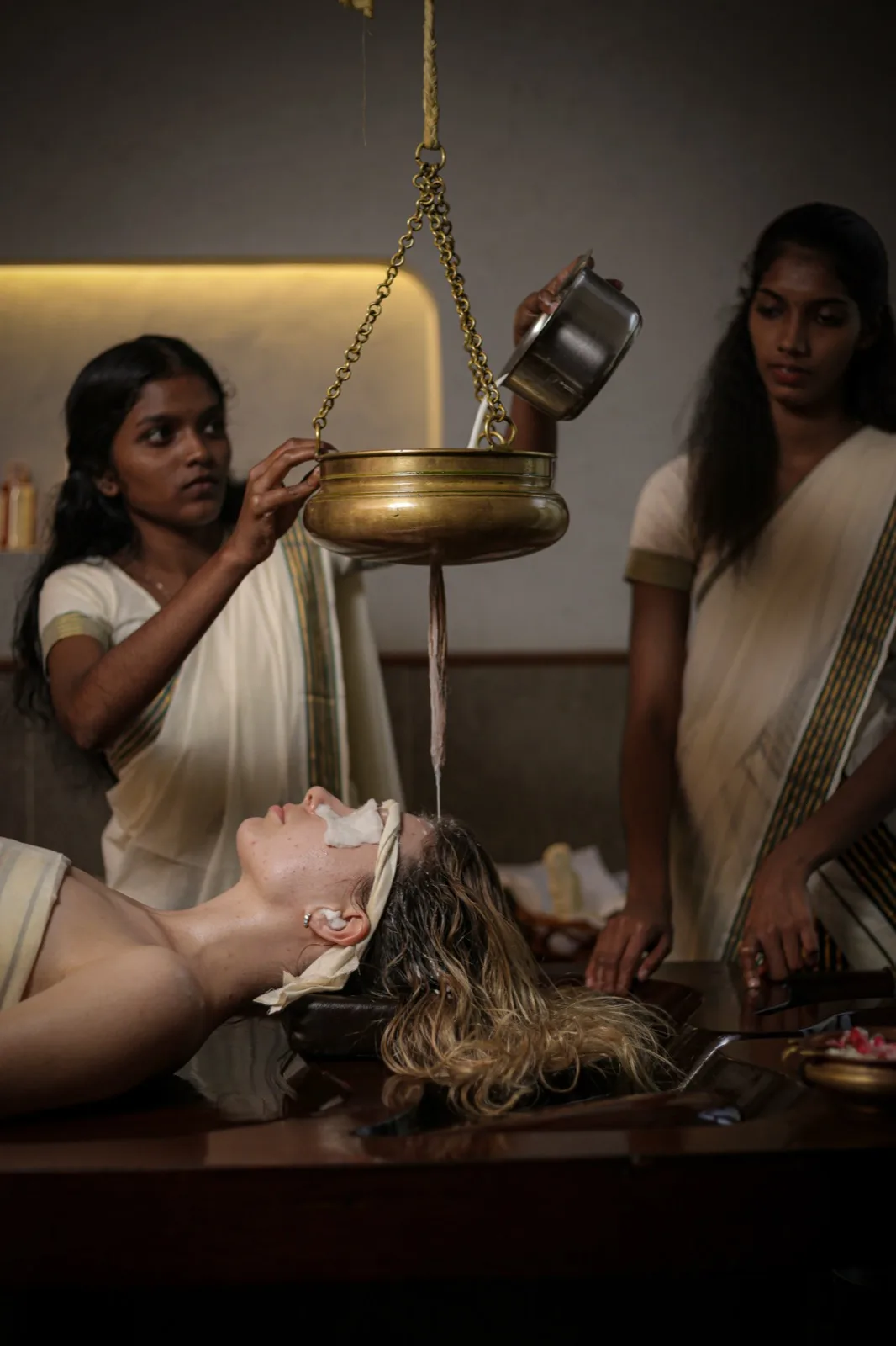 Thakaradhara: two therapists administer Thakradhara: medicated buttermilk is poured from a steel vessel into a suspended brass dhara pot streaming