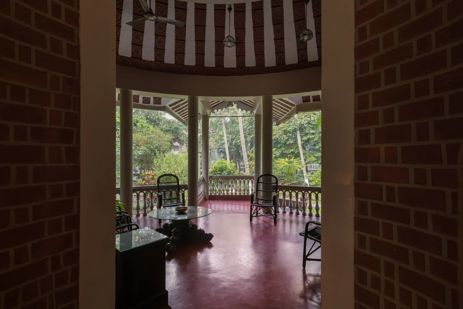 Veranda circolare aperta vista attraverso una porta in mattoni; soffitto a cupola in terracotta a strisce; sedie in canna e tavolo con piano in vetro; balaustra con vista sul giardino; pavimento in ossido marrone.