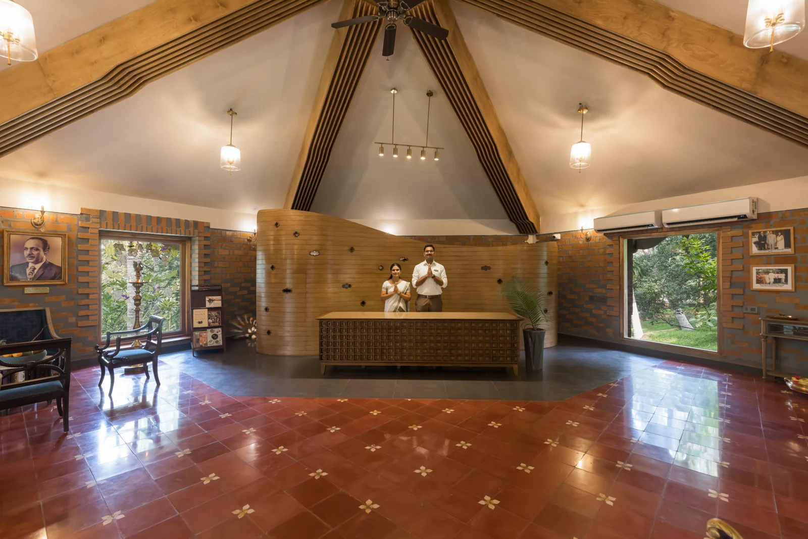 Area della reception con soffitto a volta in legno; due membri del personale in divisa che salutano a mani giunte (namaste) dietro un bancone della reception in legno intagliato; pavimento in piastrelle rosse decorate e pareti in mattoni.