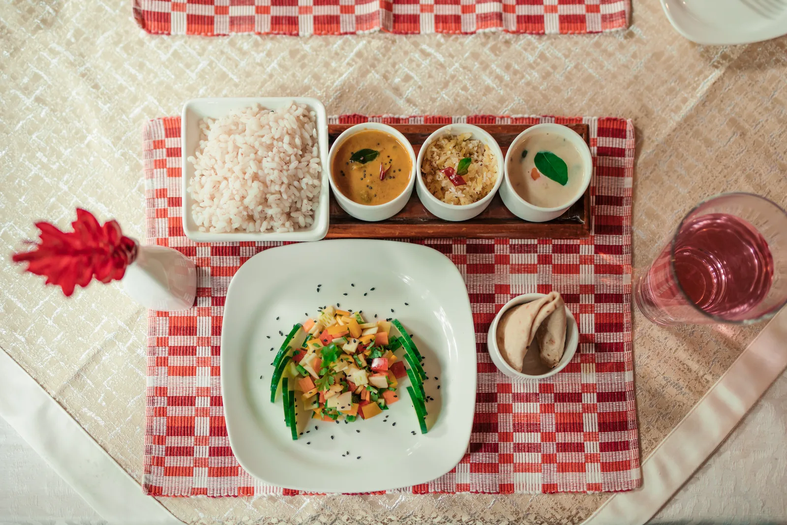 لقطة علوية لطبق ثالي نباتي: أرز أحمر؛ ثلاثة أوعية من الكاري على صينية خشبية؛ طبق من سلطة الخضار الطازجة؛ خبز تشاباتي وماء أعشاب وردي اللون على حصائر منسوجة.