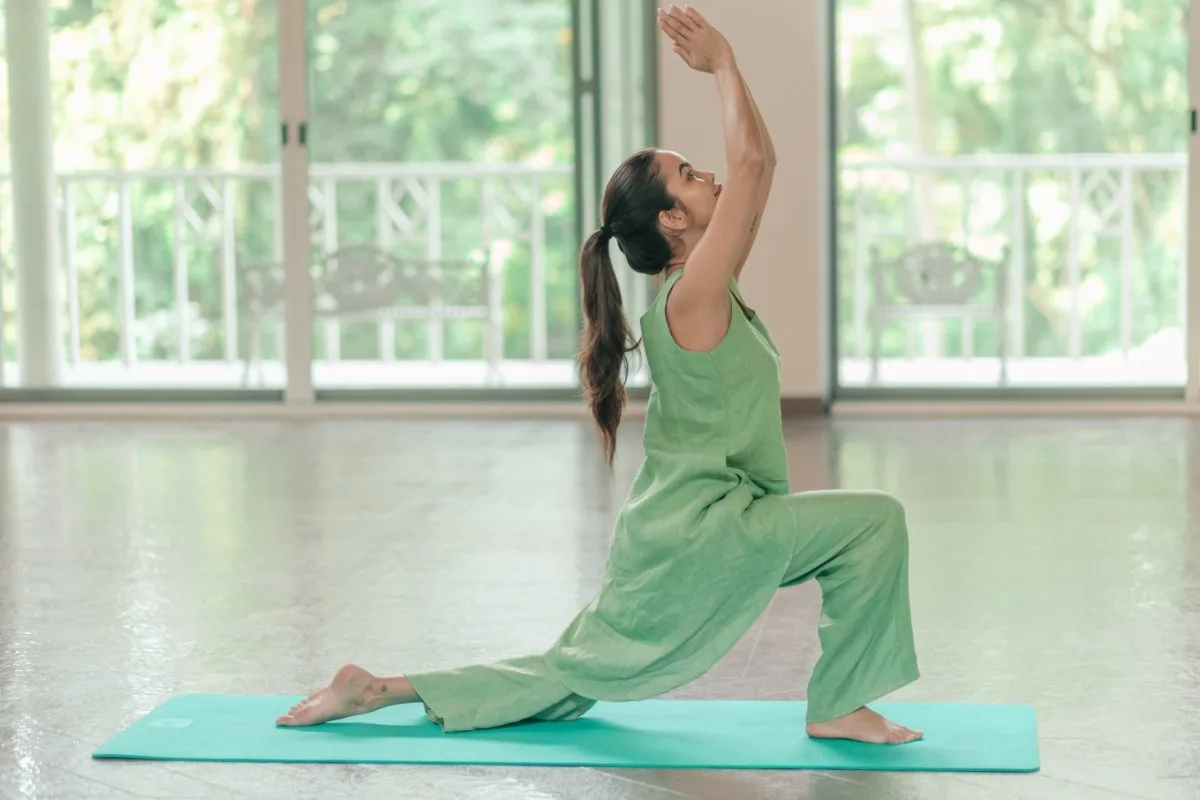 Paciente con kurta verde en posición de media luna baja sobre una esterilla turquesa, con un brazo levantado, delante de las puertas de cristal y la balaustrada blanca de la sala de yoga